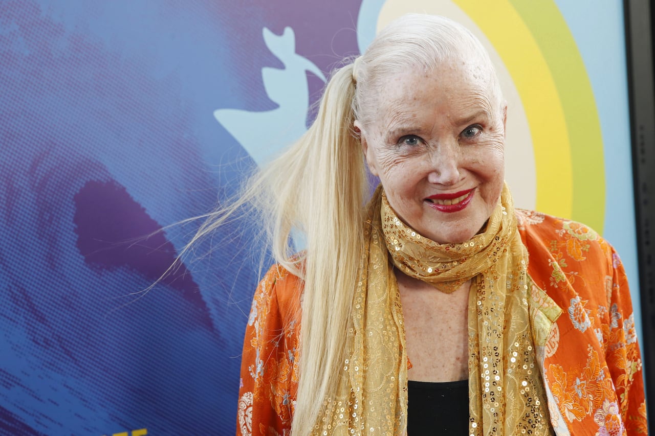 an older woman with blonde hair in a long ponytail and a beaded scarf smiles at the camera