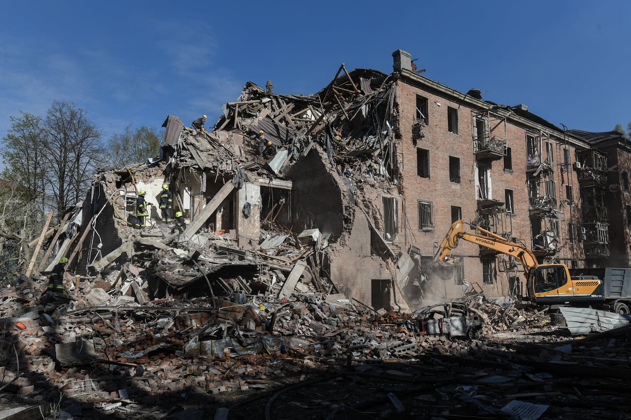 Workers clear the rubble of a large, destroyed building, a yellow bobcat style construction vehicle is on the side